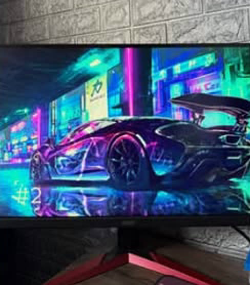 Monitor gamer ligado mostrando um carro neon, com a parede de pedra da loja ao fundo